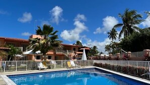 A clear blue swimming pool with a white tiled edge, surrounded by a white fence. There are lounge chairs and umbrellas near the pool, and palm trees in the background. - Recanto Sonhos do Porto (Ipojuca)