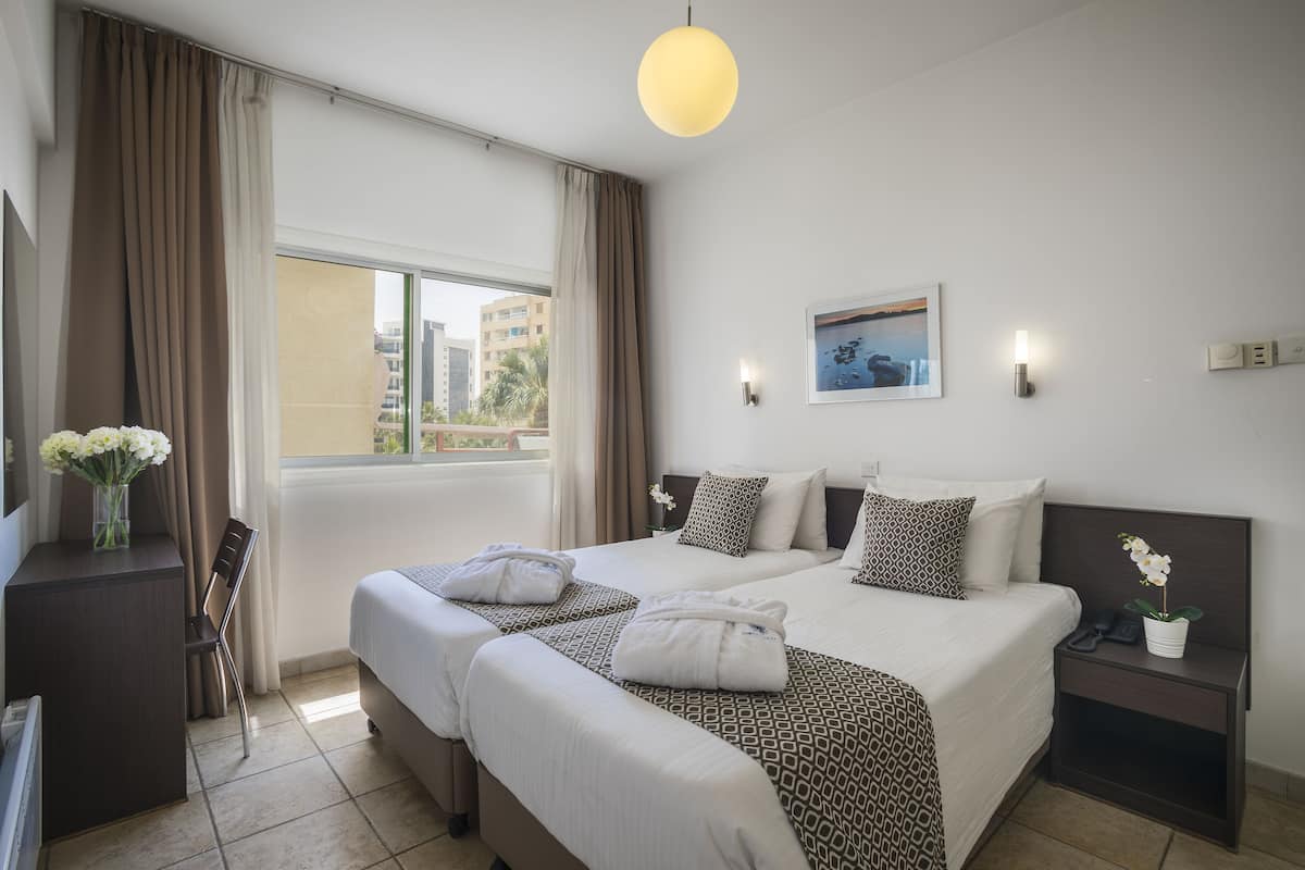 Ein Hotelzimmer mit zwei Betten, einem Schreibtisch, einem Sessel, einem Nachttisch mit Telefon und einem Fenster mit Vorhängen.