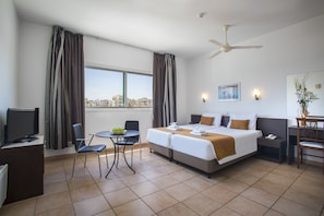 A hotel room with a bed, a desk with a computer, a chair, a small table with a bowl of fruit, and a window with curtains.
