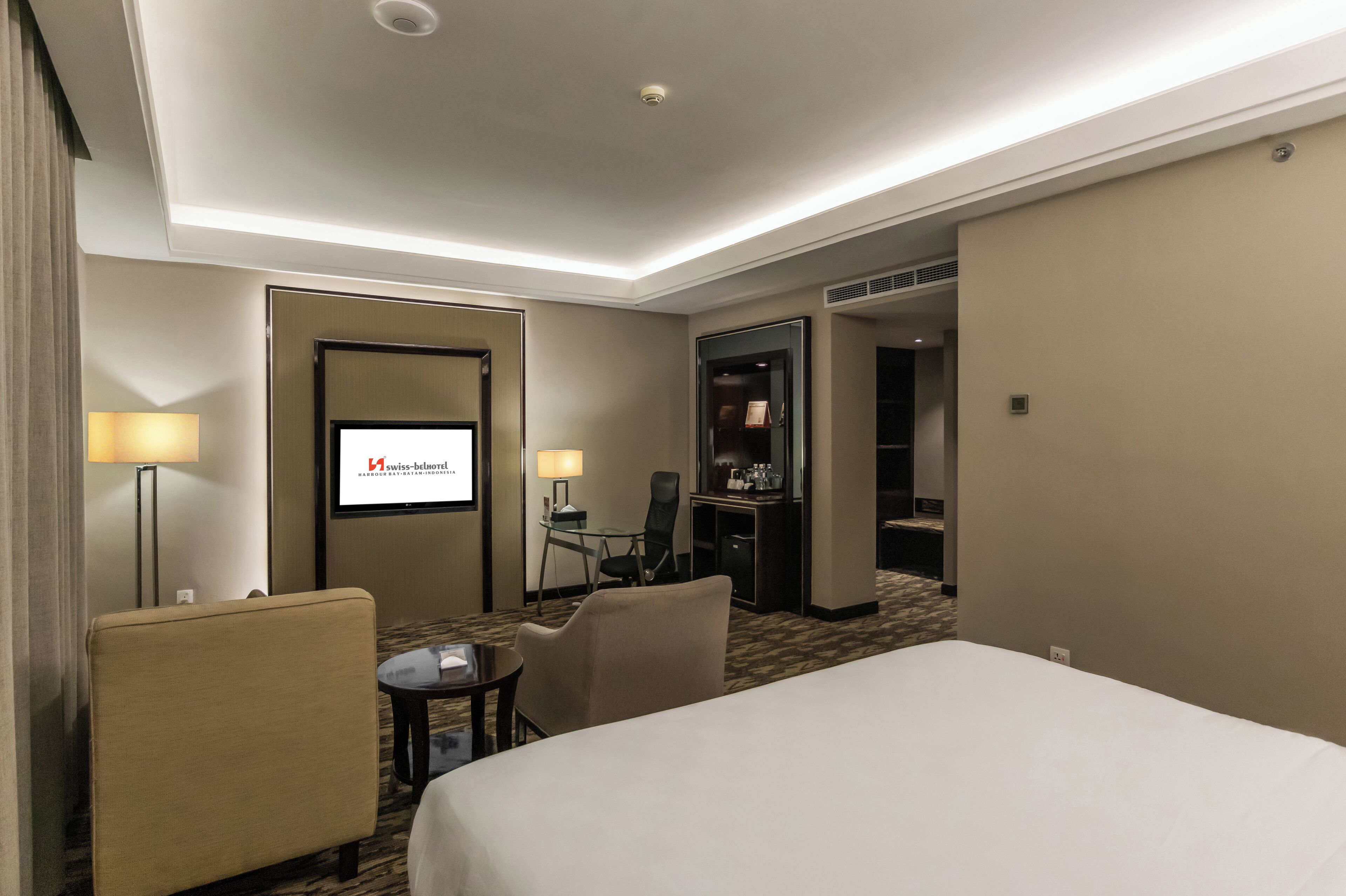 A hotel room with a bed, a desk, a chair, a TV screen, and a minibar.