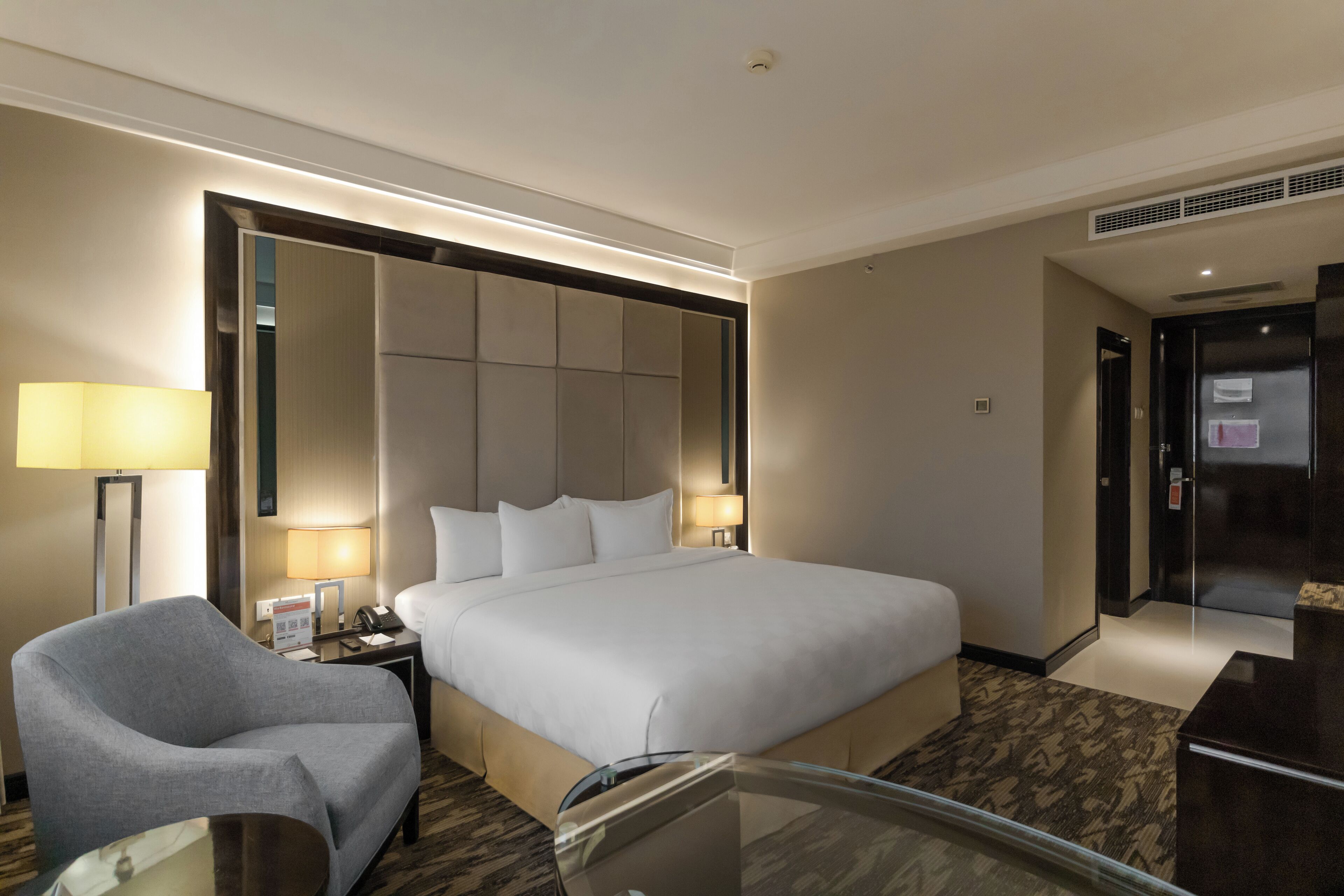 Habitación de hotel con una cama grande, una mesita de noche con lámpara, una silla y una mesa de vidrio.