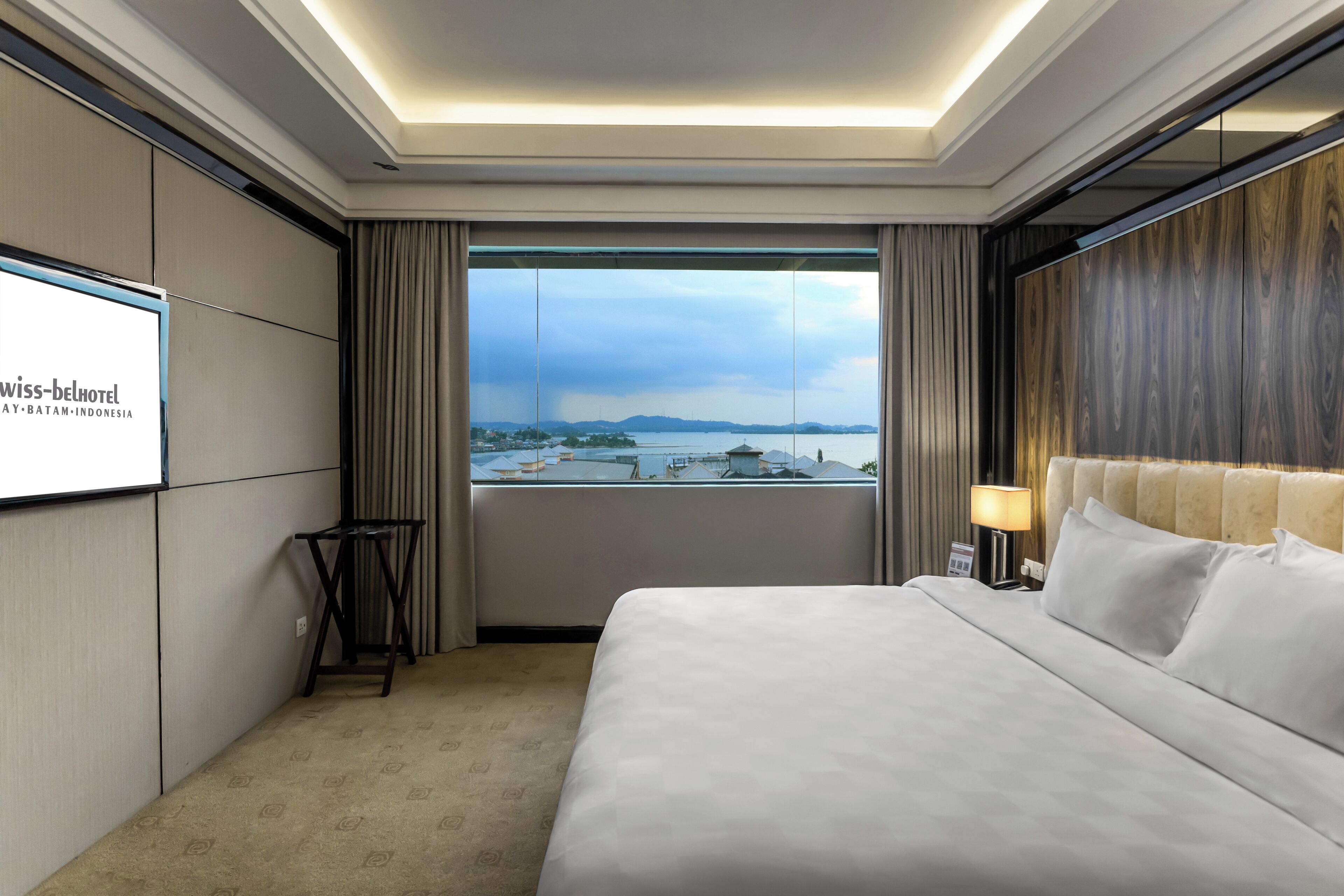 A hotel room with a large bed, a TV screen, a view of the city, and a window with curtains.