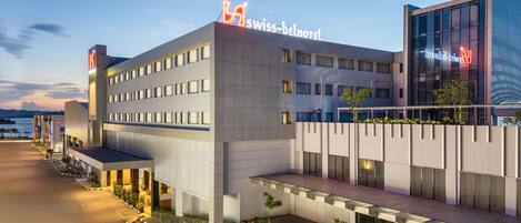 A modern hotel with a prominent sign reading "Swiss-Belhotel." It features a flat roof, multiple windows, and a well-lit entrance area.