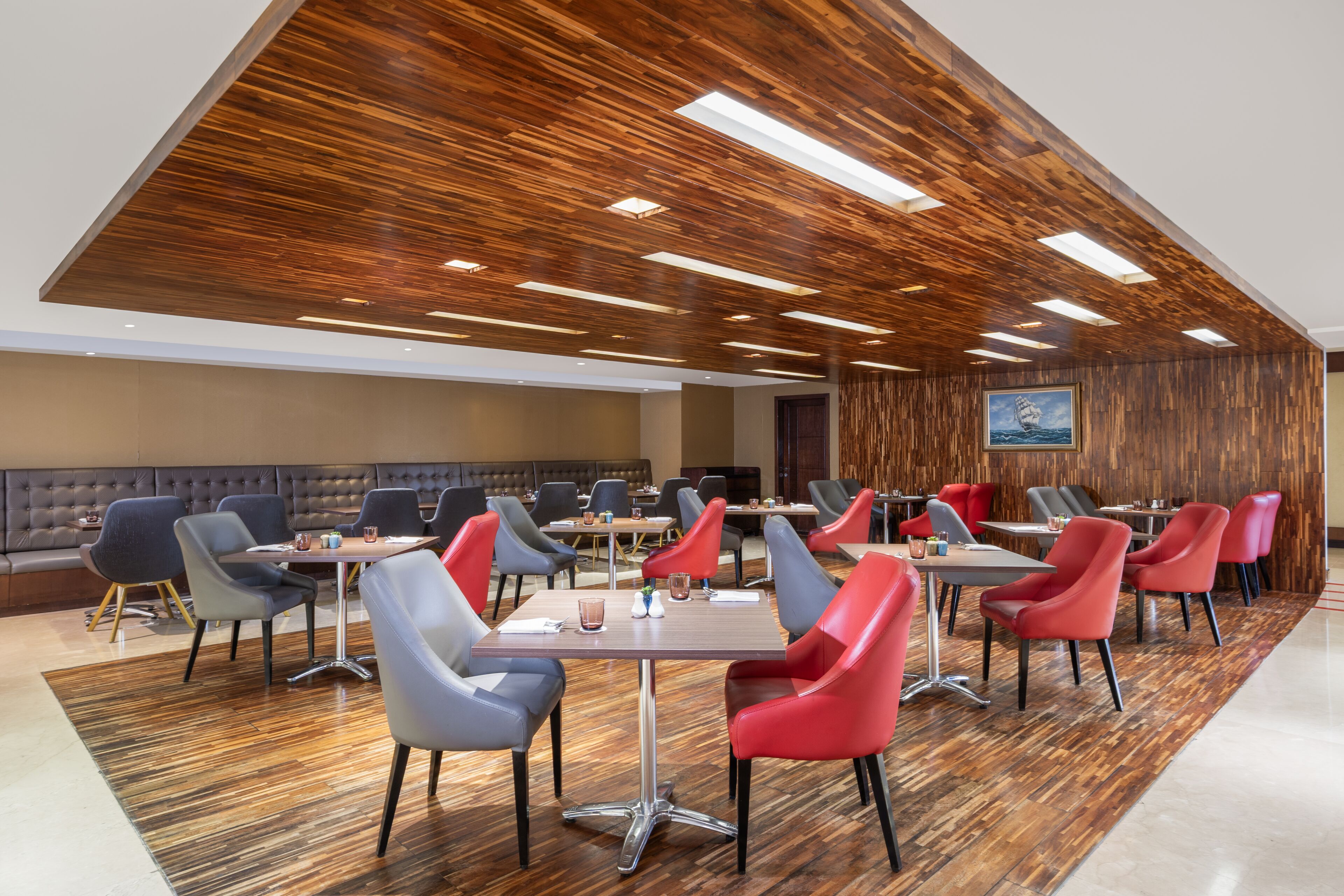 A modern dining area with wooden ceiling panels, multiple tables and chairs, and a large painting on the wall.