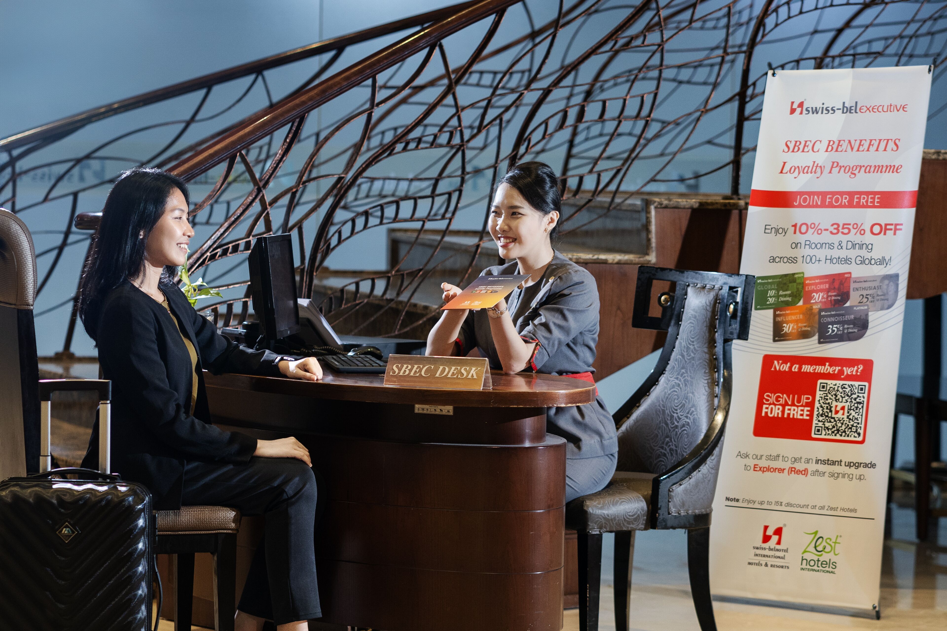 A reception desk with a sign for the Swiss-Bel-executive SBE benefits loyalty program, a QR code for sign-up, and a promotional offer for discounts on rooms and dining.