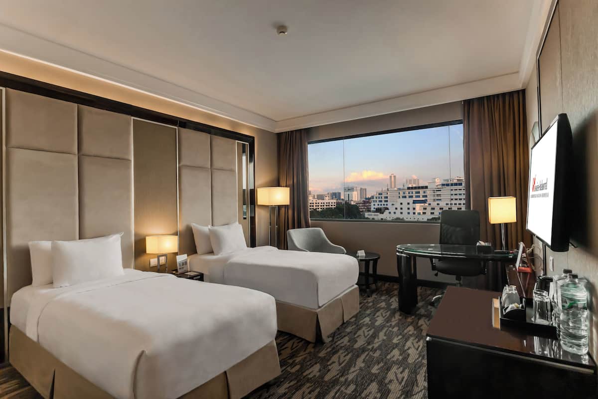 Een hotelkamer met twee bedden, een bureau, een stoel, een televisie en een groot raam met uitzicht op de stad.