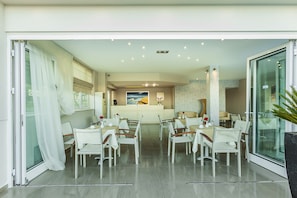 Zona pranzo moderna con sedie e tavoli bianchi, una reception e vista sul mare attraverso un'ampia finestra.