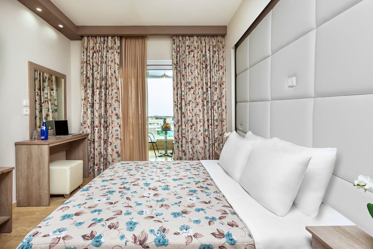 Una camera d'albergo con un letto, una scrivania e una sedia. Si vede una finestra con tende, e un balcone è visibile attraverso la finestra.