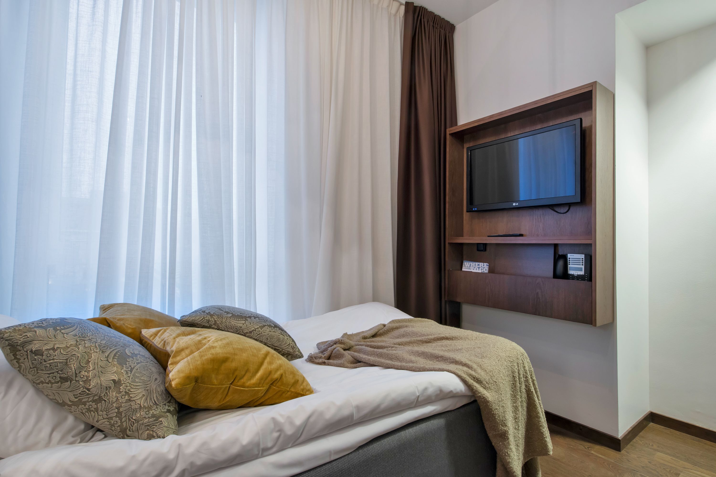 Une chambre d’hôtel avec un lit, un téléviseur fixé au mur et une fenêtre avec des rideaux légers.