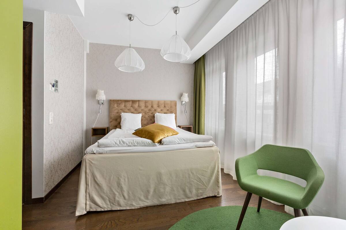 A modern hotel room with a large bed, a green chair, and a view of the city.