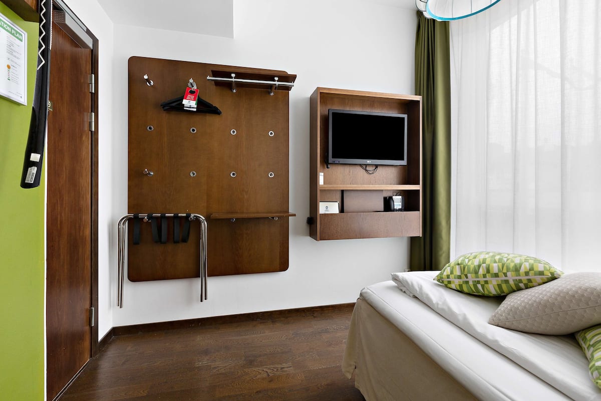A hotel room with a wooden wardrobe, a mounted TV, a bed with pillows, and a green curtain.
