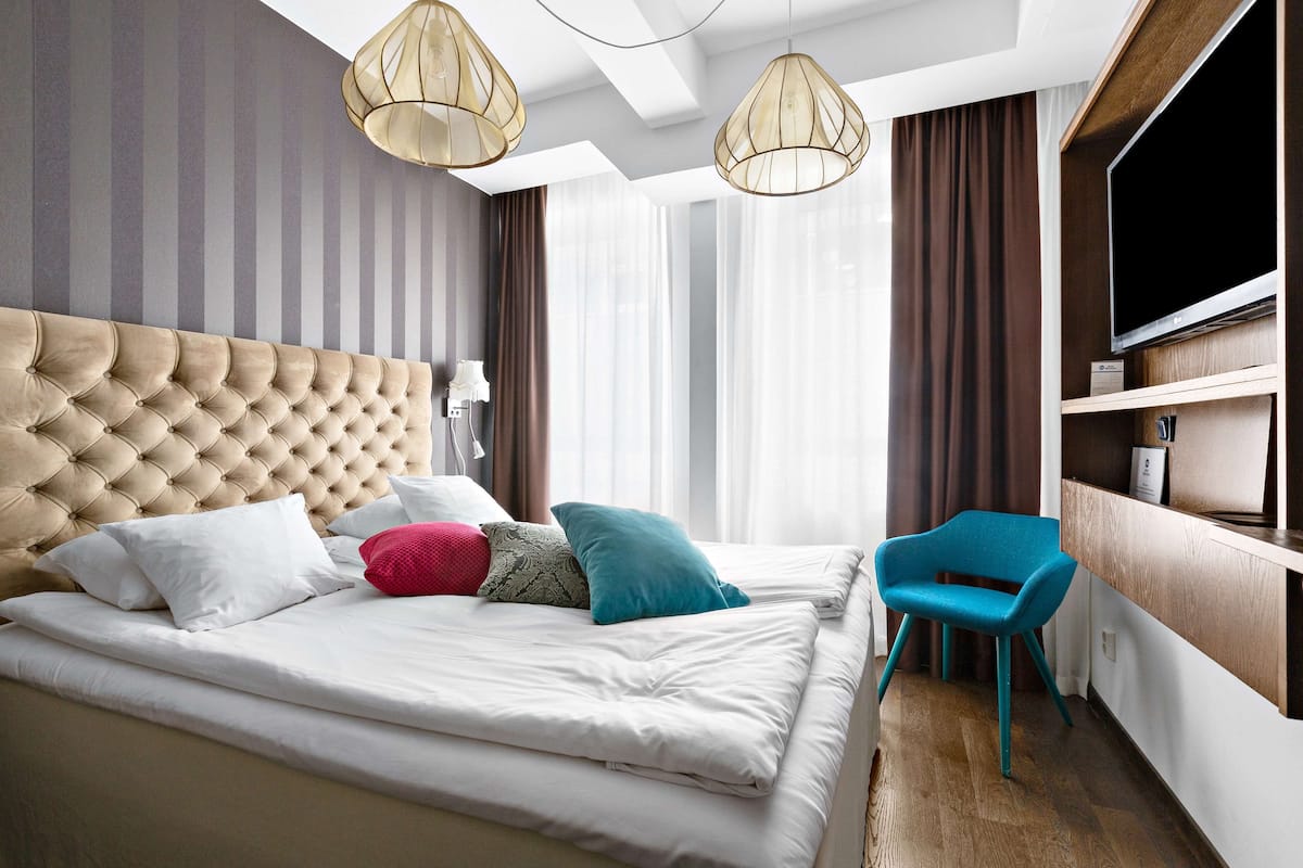A modern hotel room with a tufted headboard, a bed with white linens, a teal chair, and a flat-screen TV on a wooden shelf.