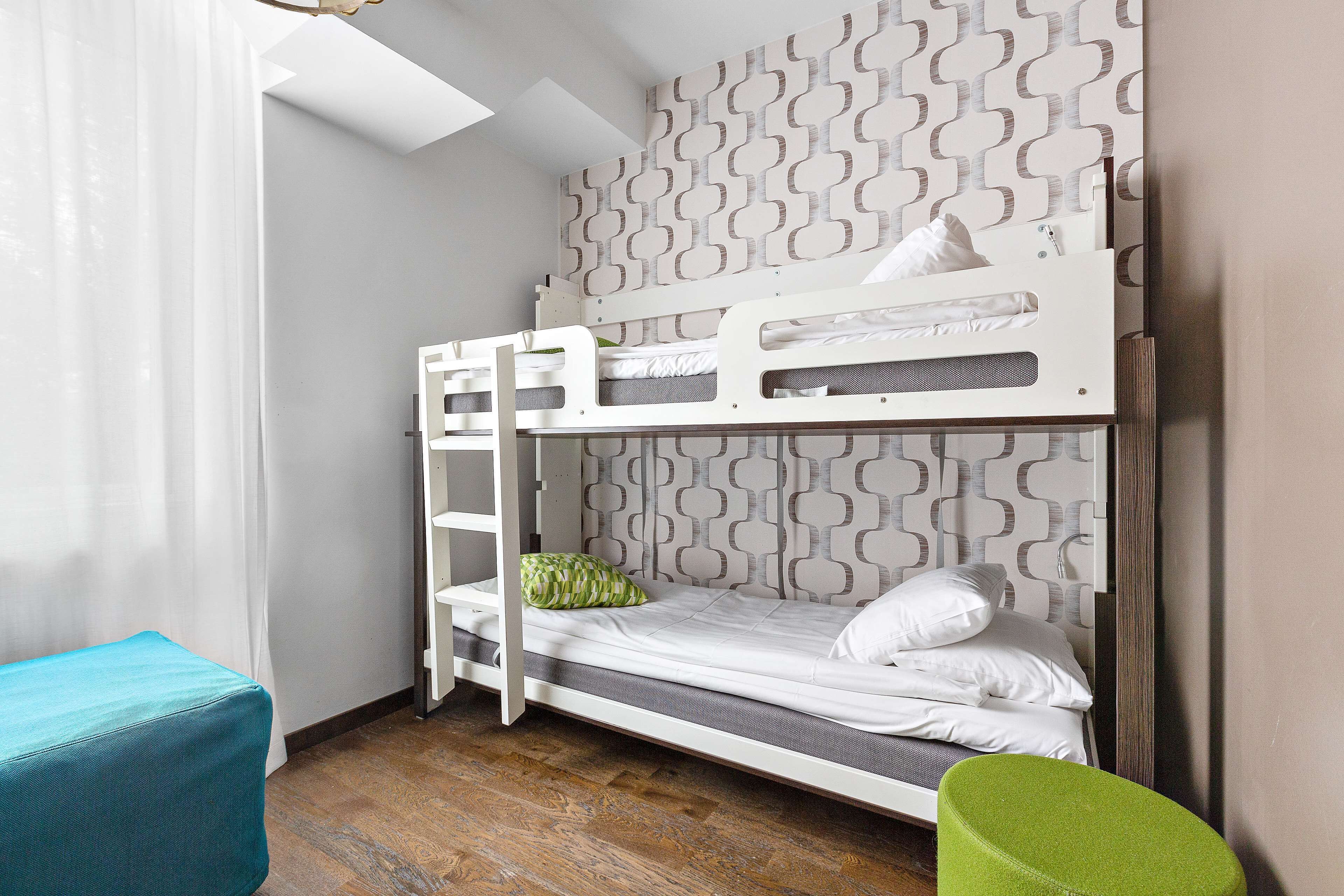 Une chambre avec un lit superposé, un mur décoré de motifs, un plancher en bois et un pouf turquoise.