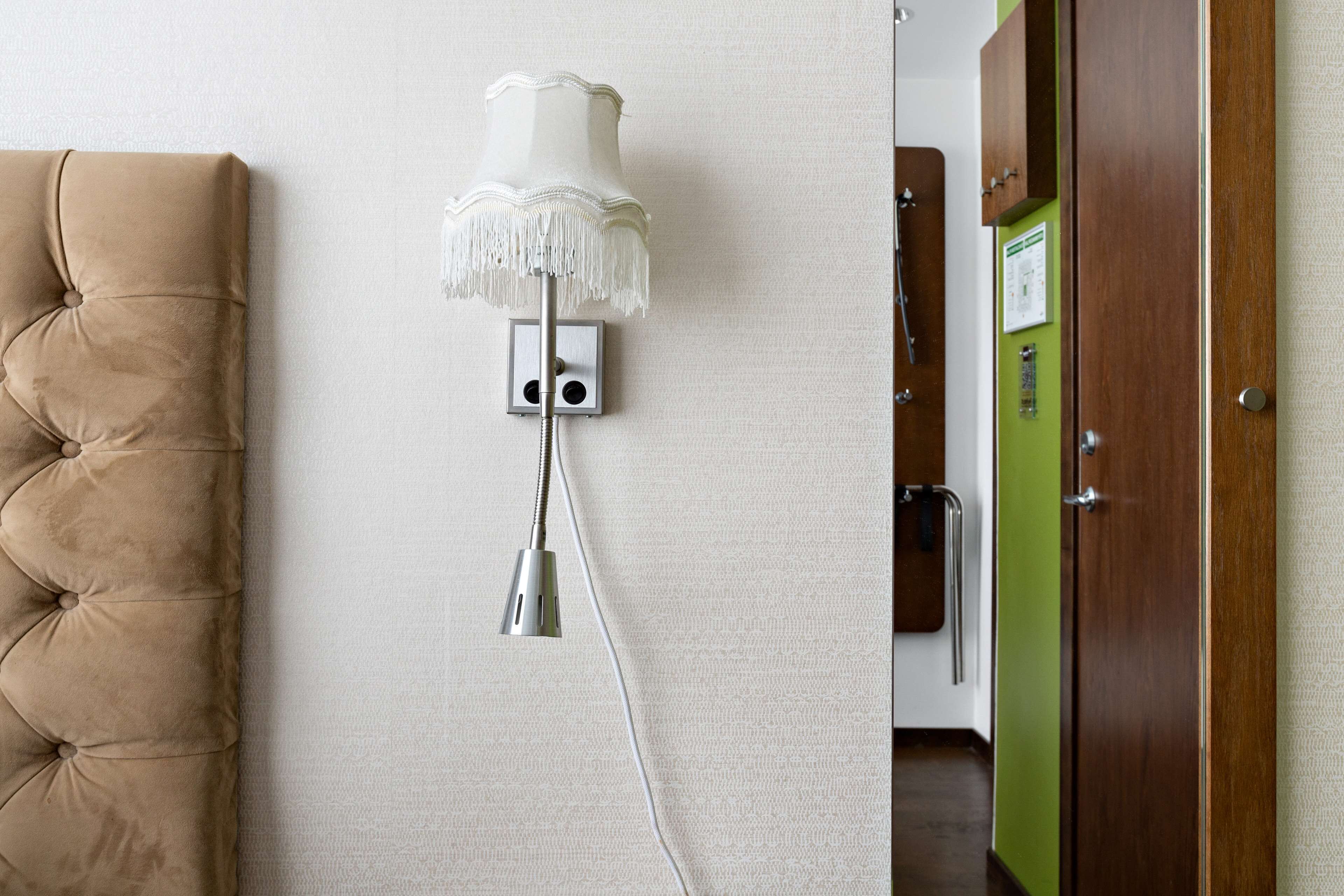 Una lámpara de pared con pantalla blanca y base metálica, un cabecero marrón capitonado y una puerta verde al fondo.