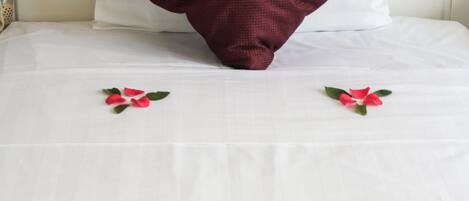 Un lit recouvert d’une couverture blanche, un oreiller rouge foncé et une composition en forme de cœur faite de pétales de roses rouges.