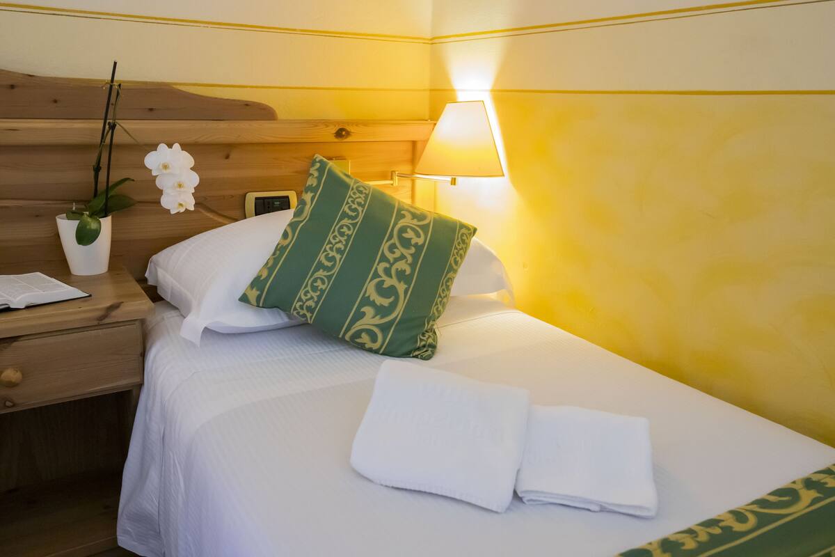 A neatly made bed with white linens, a green patterned pillow, and a white towel folded at the foot of the bed. A small white flower pot with a single white orchid is placed on the nightstand beside the bed.
