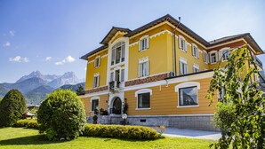 A yellow building with a balcony, surrounded by green lawns and mountains in the background. - Villa Imperina (Agordo)