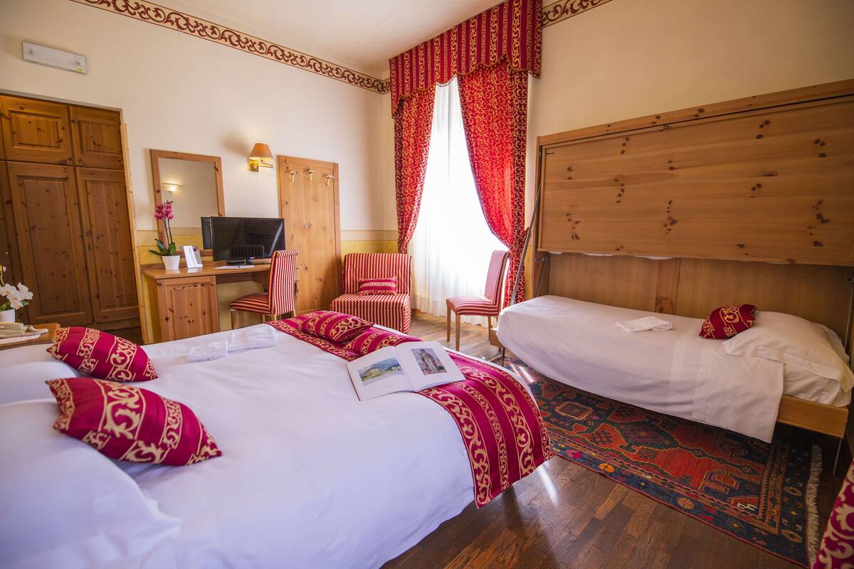 A hotel room with two beds, a wooden wardrobe, a desk with a TV, and a large window with red curtains.