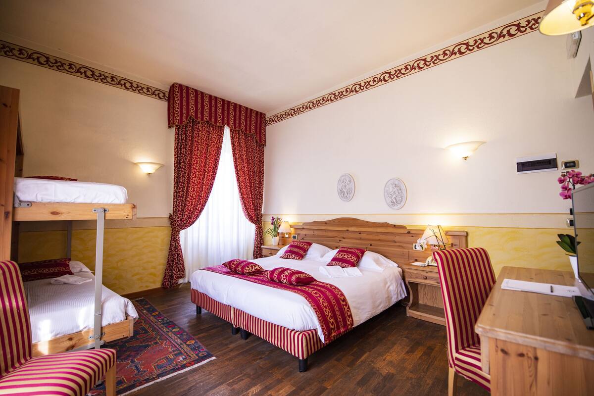 A room with a bunk bed, a single bed with red and white bedding, a wooden desk, a chair, and a nightstand with a lamp.