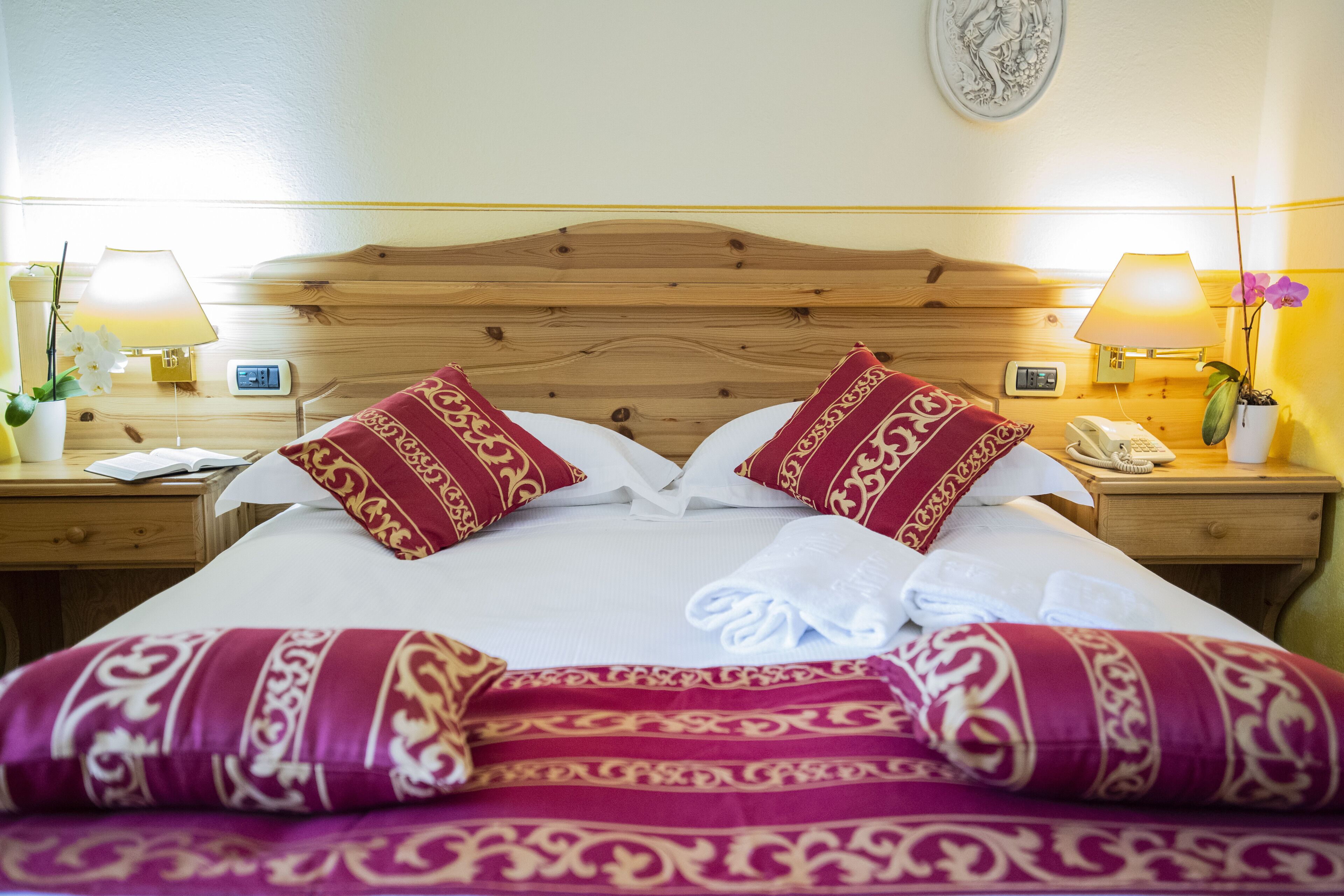 Een net opgemaakt bed met rode en gouden kussens, een houten hoofdbord, bedlampen en een kleine plant in een pot.