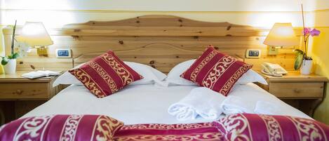 A neatly made bed with red and gold patterned pillows, a wooden headboard, bedside lamps, and a small potted plant.