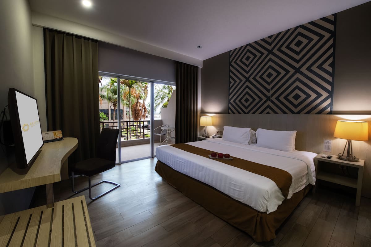 Ein Hotelzimmer mit einem großen Bett, zwei Nachttischen inklusive Lampen, einem Schreibtisch mit Stuhl und Blick auf Palmen durch eine Schiebeglastür.