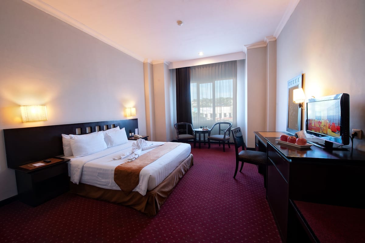 Ein Hotelzimmer mit einem großen Bett, Nachttischen, einem Schreibtisch mit Fernseher und Blick ins Freie durch ein Fenster.