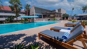 Un espace aménagé au bord de la piscine, avec des chaises longues et des parasols, une piscine à l’eau d’un bleu limpide, et un bâtiment avec un toit recouvert de tuiles rouges en arrière-plan.