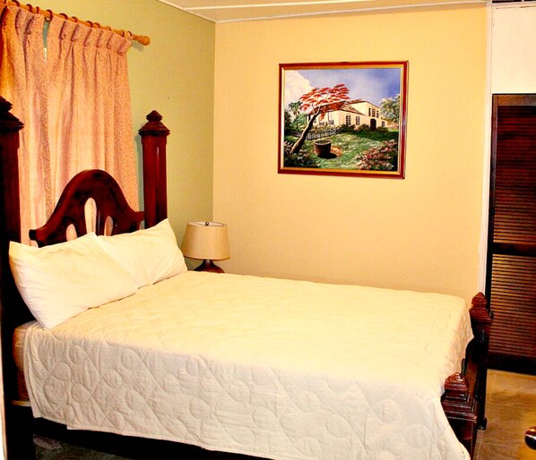 A neatly made bed with a quilted cover, a wooden headboard, a bedside lamp, and a framed painting of a landscape on the wall.