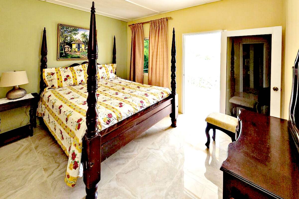 Una cama con dosel adornada con un edredón floral, una mesita de noche con una lámpara, un banco de madera y una puerta que conduce a otra habitación.