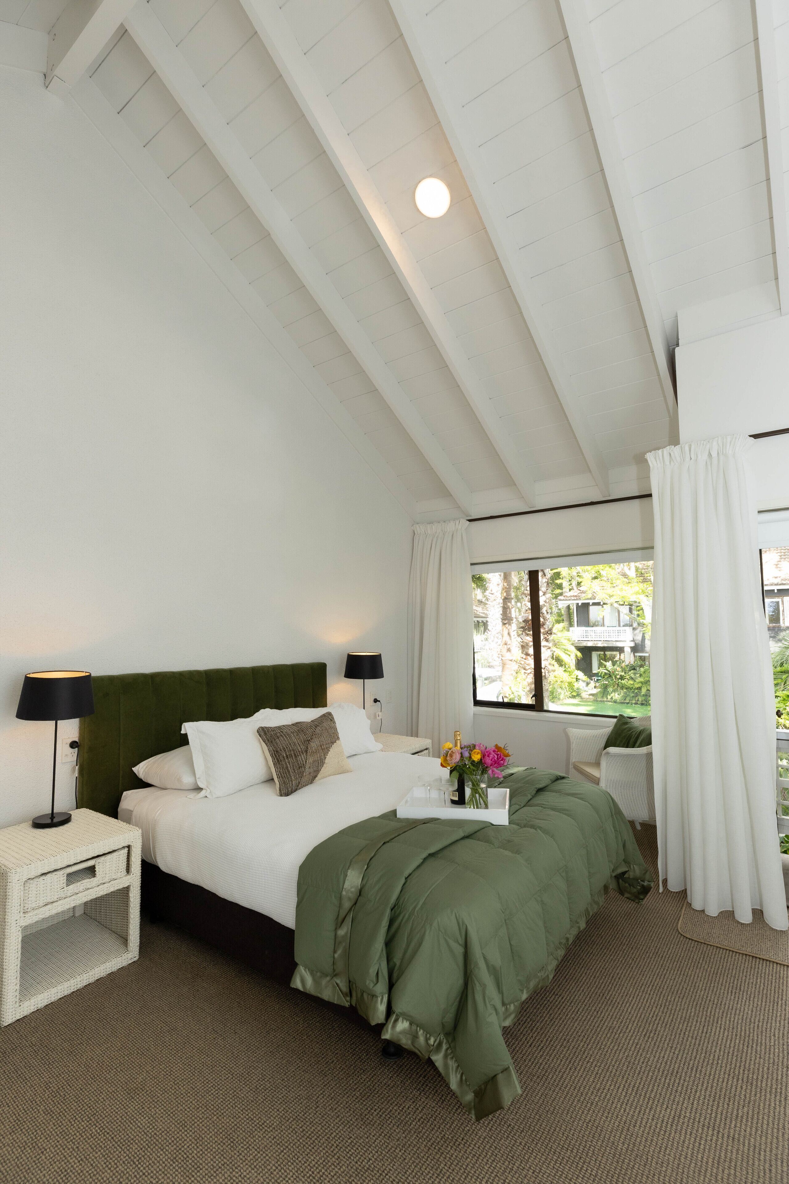Een slaapkamer met een groen bed, witte wanden, een raam met gordijnen en een nachtkastje met een lamp.