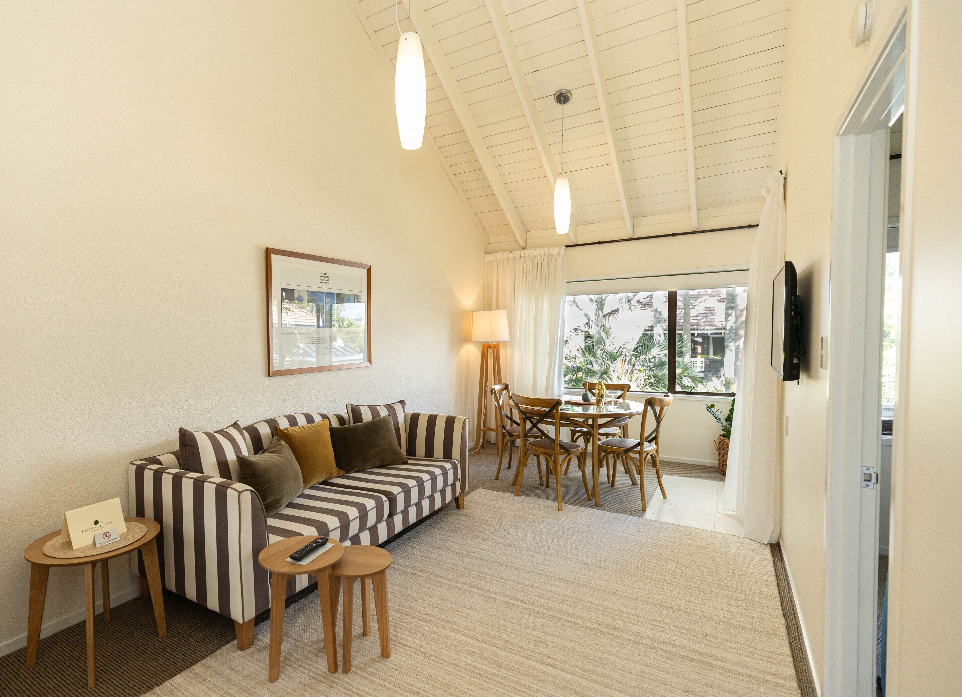 A cozy living room with a striped sofa, a small table with a remote, a framed picture on the wall, and a dining area with chairs and a table.