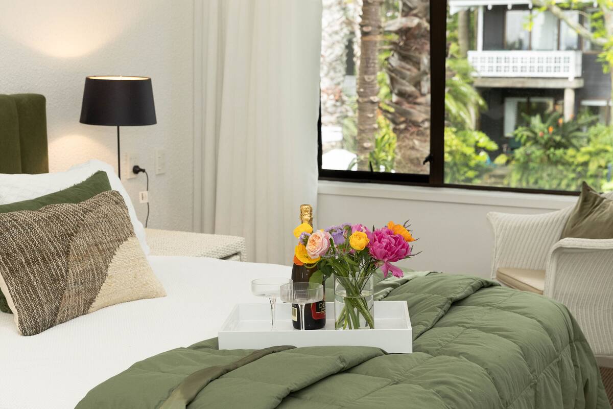 Una cama bien hecha con un edredón verde, una mesita de noche con una botella y flores, y una vista de un edificio a través de una ventana.