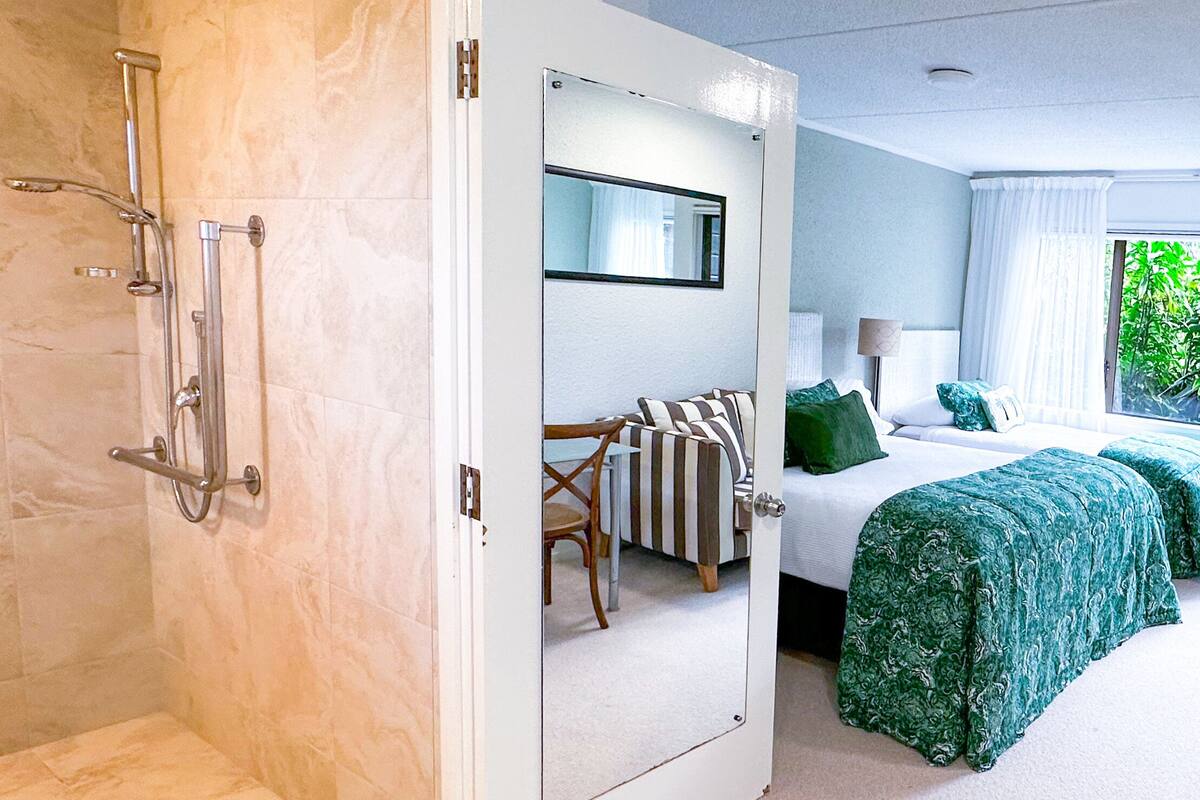 Habitación de hotel con zona de ducha, cama con ropa de cama verde y un espejo grande.