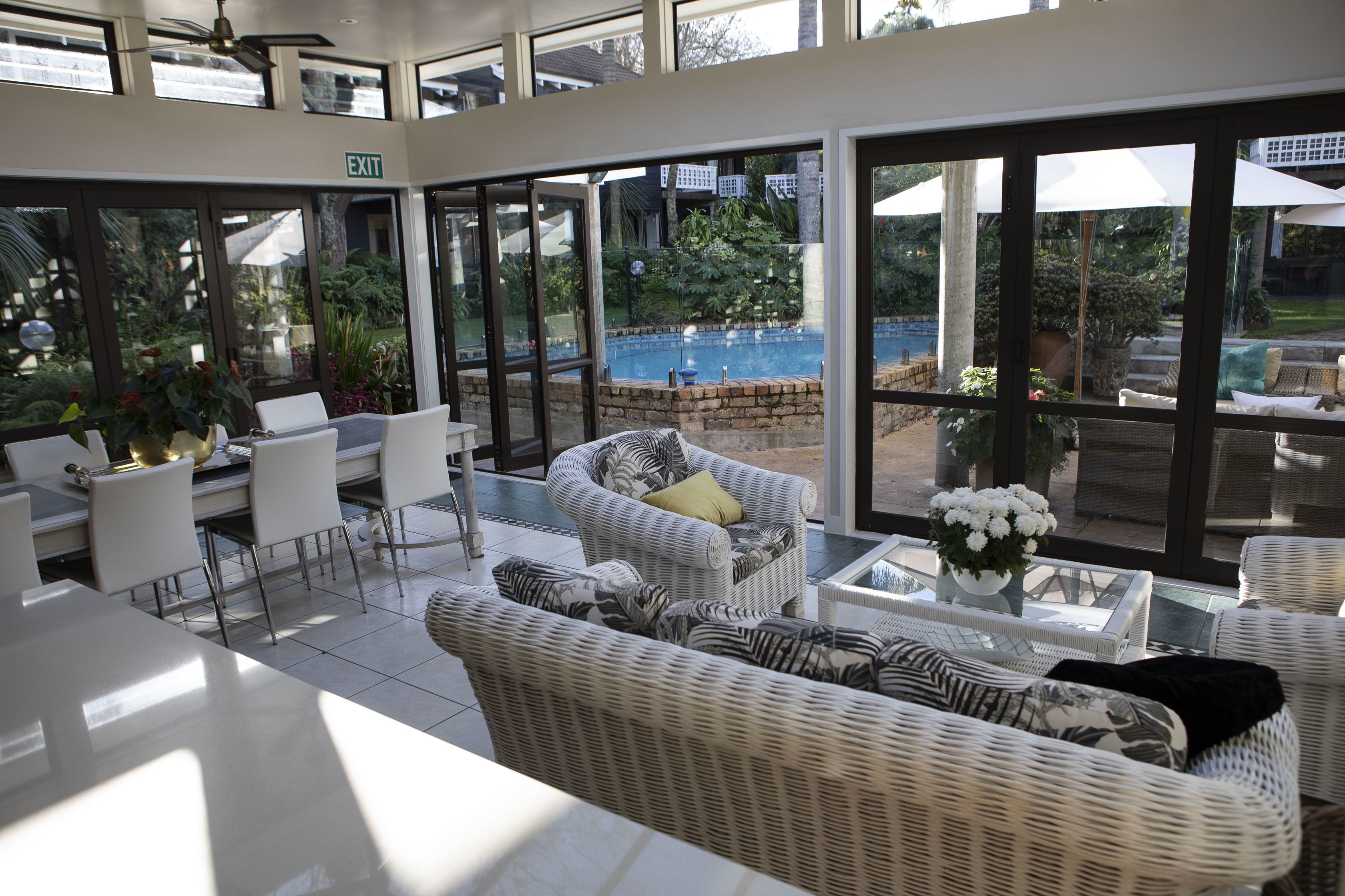 Un espace intérieur lumineux, baigné de soleil, avec des meubles en osier, une vue sur la piscine et un panneau indiquant la sortie.