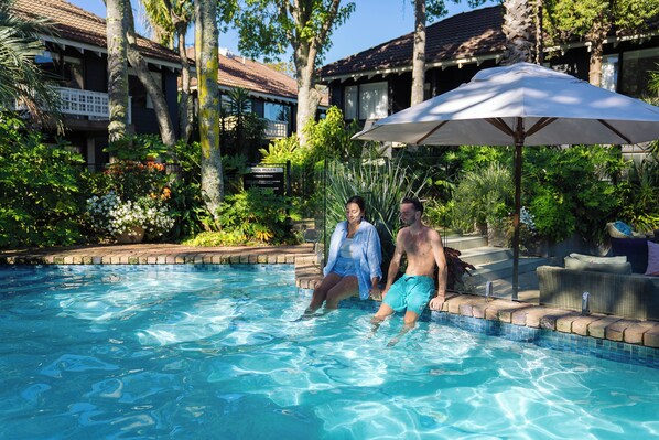 A swimming pool with a couple sitting on the edge, surrounded by lush greenery and a building with a balcony.