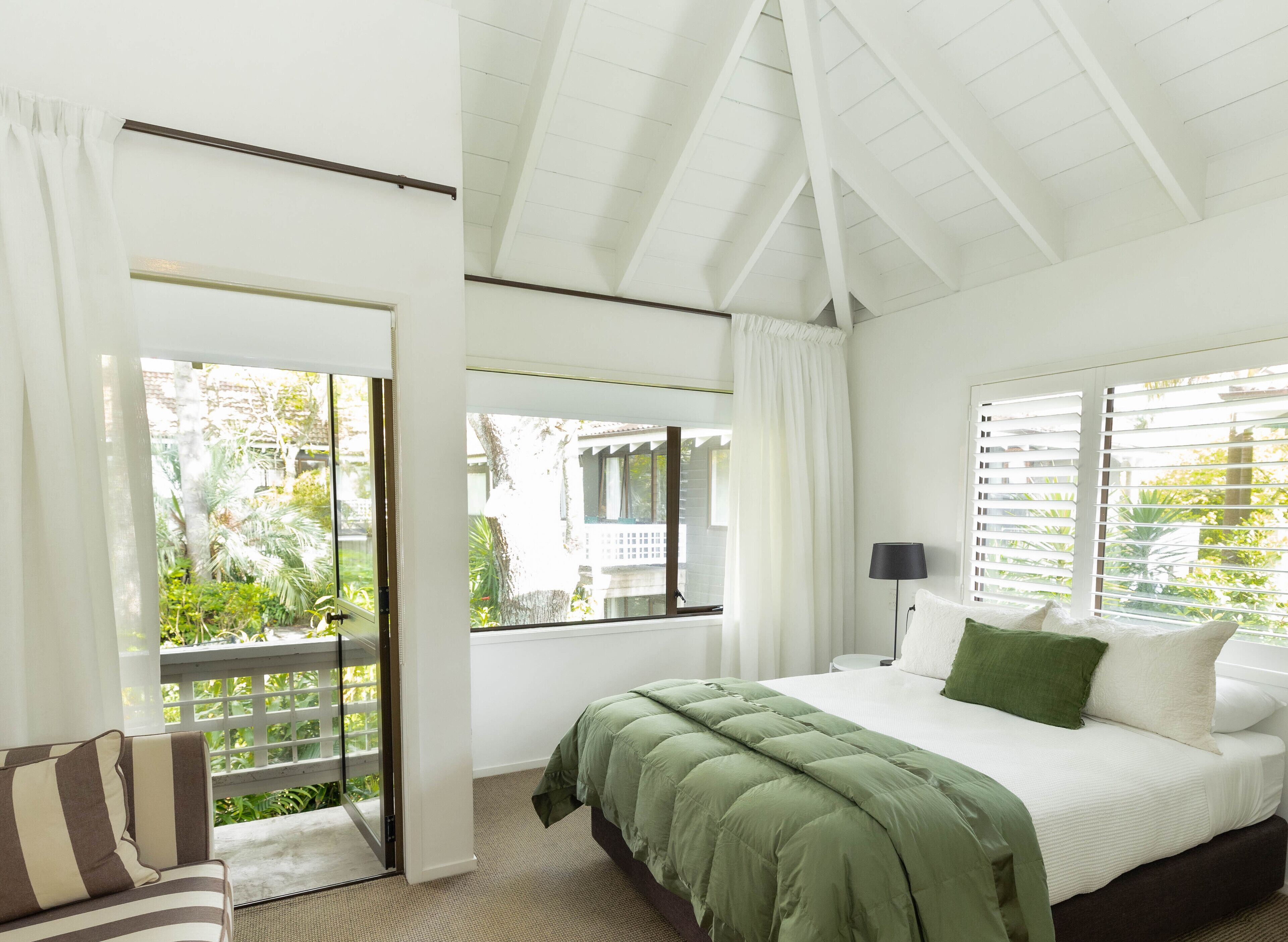 A bedroom with a bed, a nightstand, a lamp, and a view of greenery outside.