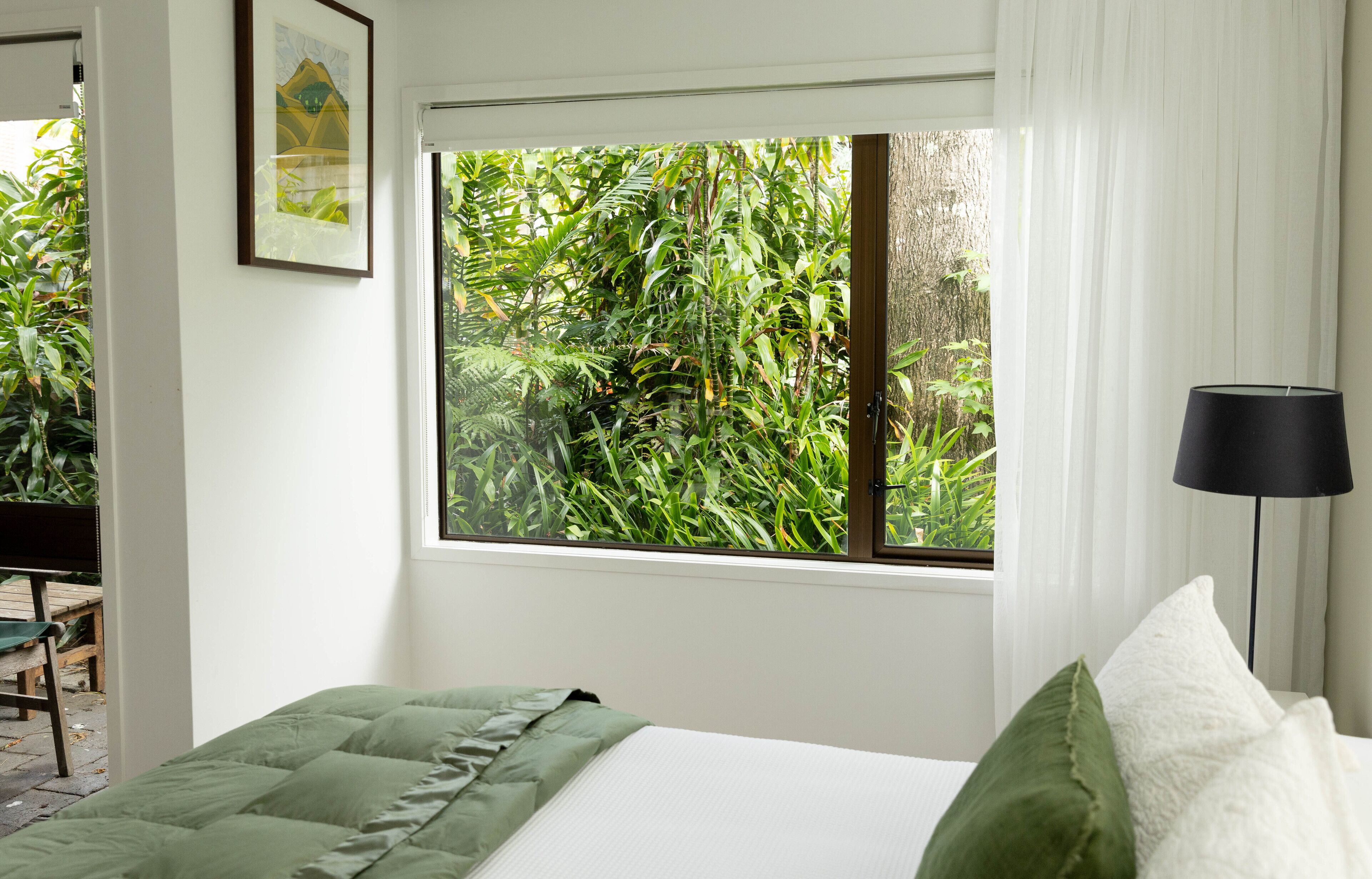 Un dormitorio con un ventanal grande, una cama con ropa de cama verde y blanca, una lámpara de pie negra y un cuadro en la pared.