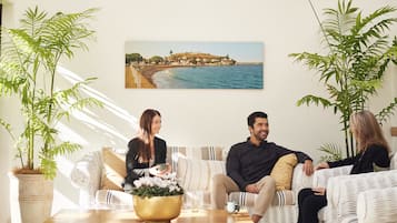 A modern living room with a sofa, two potted plants, a coffee table, and a painting of a coastal scene.