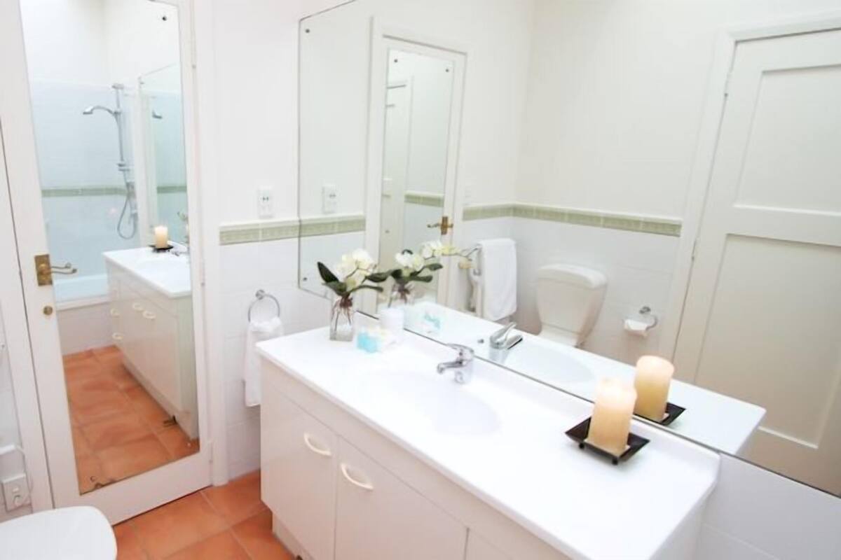 Un baño con lavamanos blanco, espejo y ducha visible a través de una puerta de cristal.