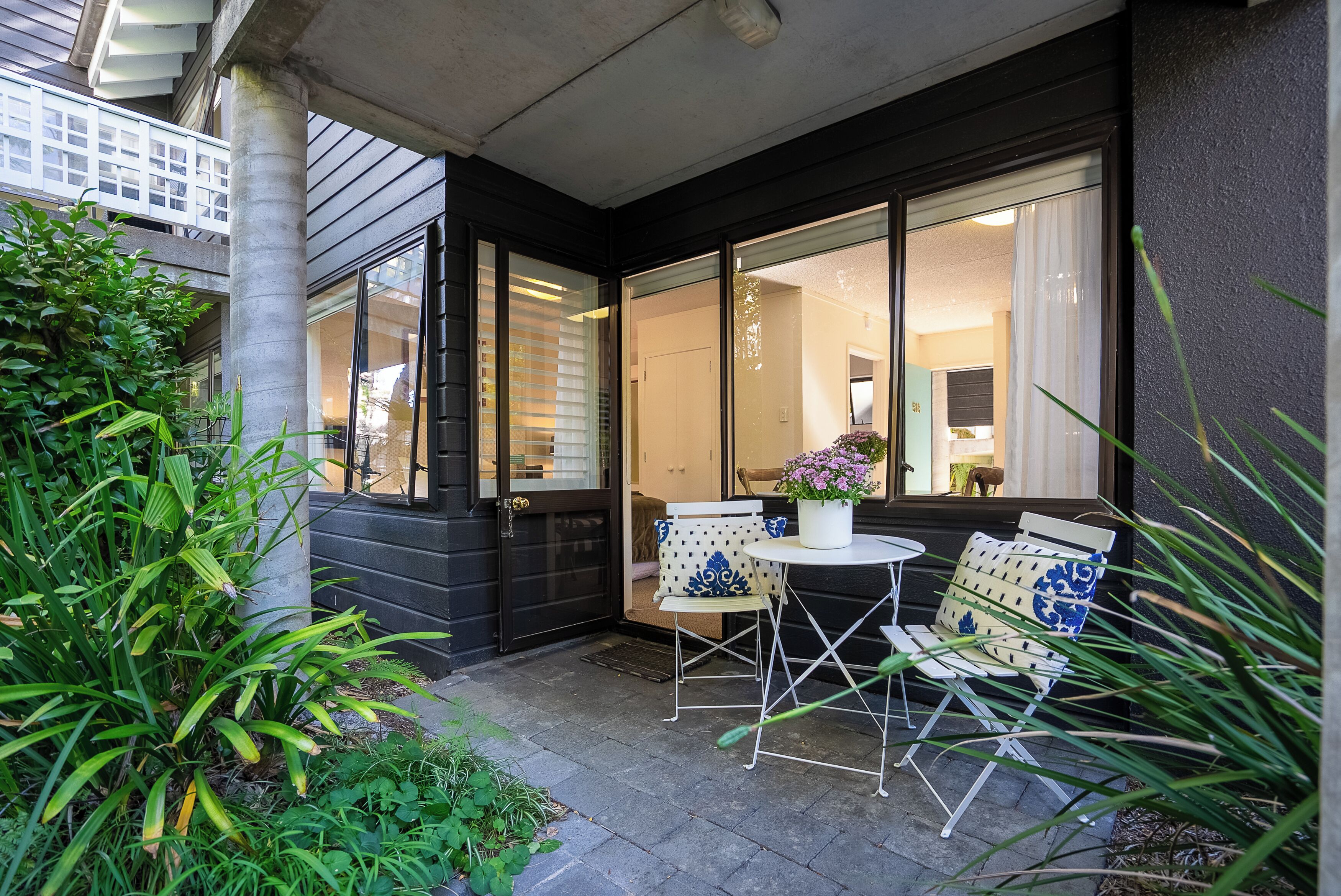 A patio with two white chairs and a table, surrounded by greenery and large windows.