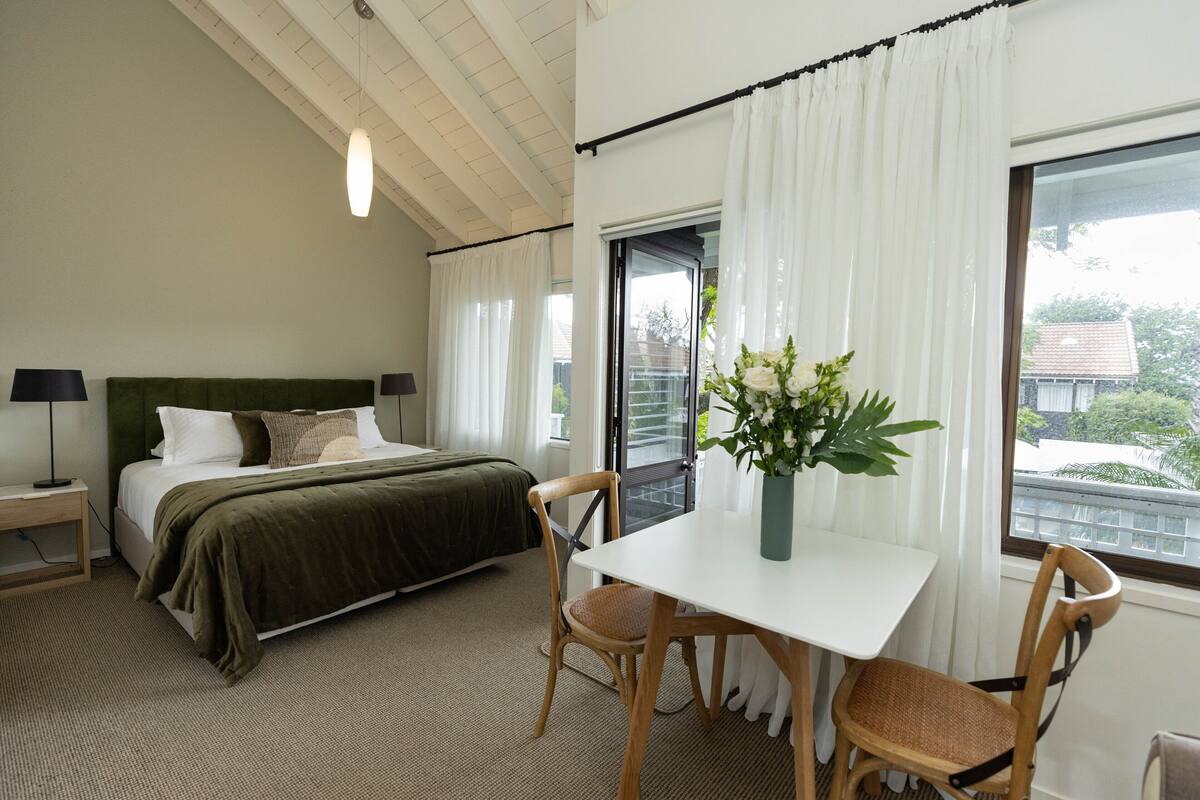 Un dormitorio con cama, una mesa, sillas, una mesita de noche y un ventanal con cortinas.