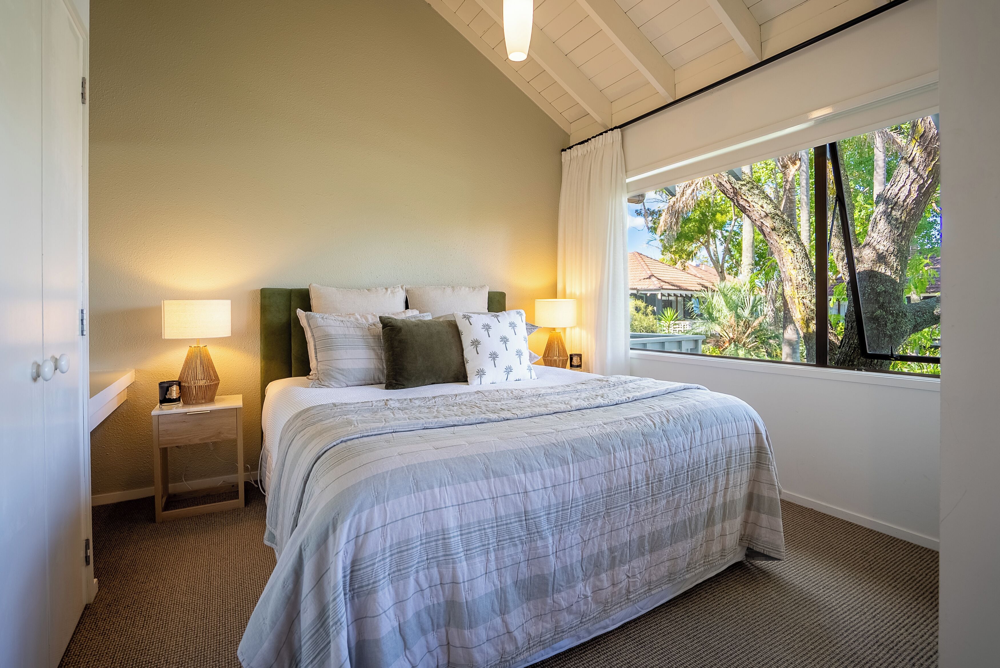 A bedroom with a large bed, two bedside tables with lamps, a view of trees and a house through a window.