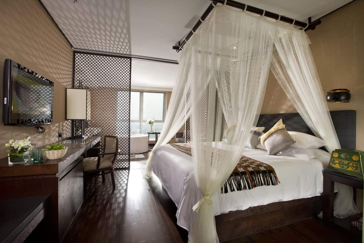 A bedroom with a four-poster bed, a desk with a chair, a television, and a window with a view.