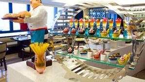 Un interno di ristorante moderno con una statua dello chef, una vetrina in vetro con dessert colorati e una vista del cielo attraverso ampie finestre.