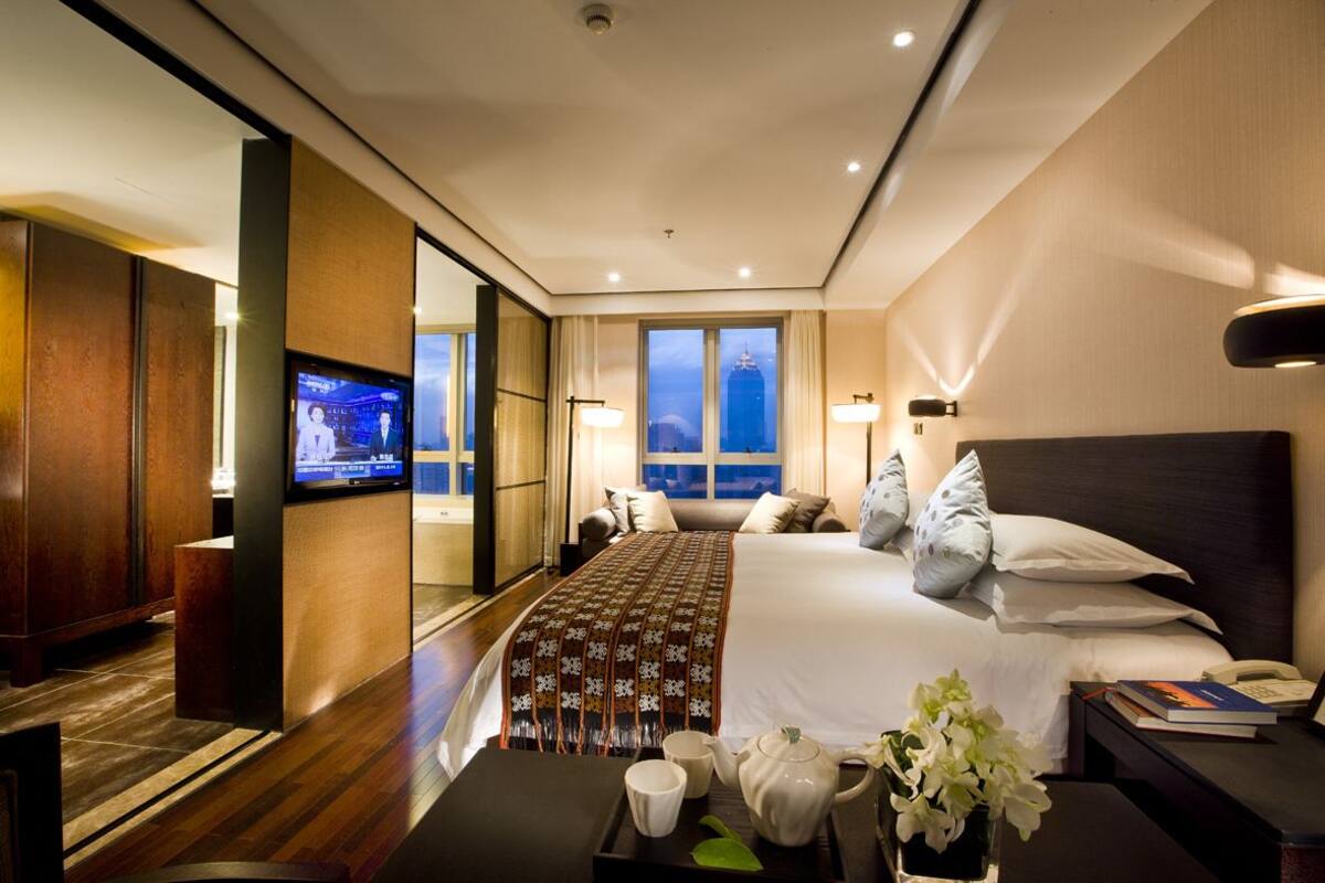 A hotel room with a large bed, a television, a desk with a phone, and a view of the city skyline through the window.