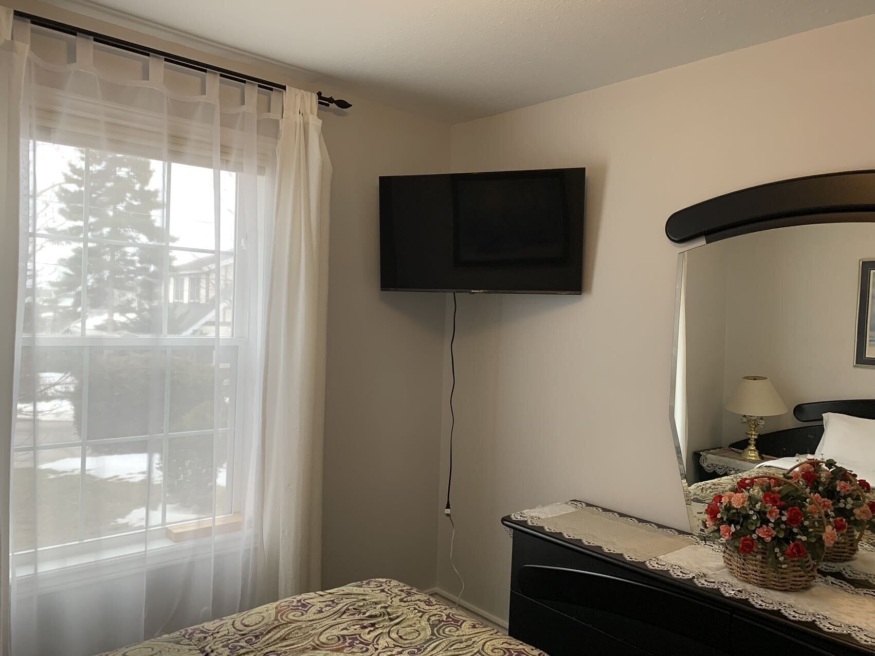 Une chambre d’hôtel avec un lit, un téléviseur fixé au mur, une table de chevet avec une lampe et une fenêtre avec des rideaux légers.
