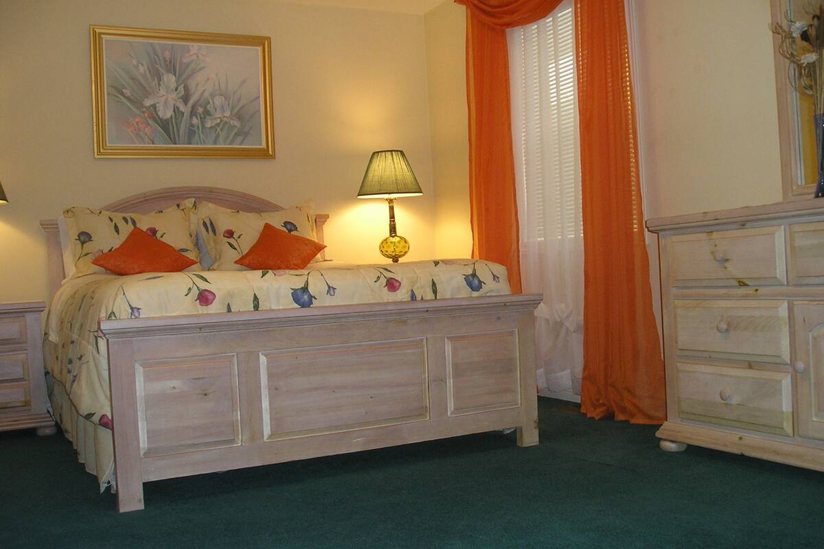 Un dormitorio con cama, mesita de noche, cómoda, lámpara y un cuadro en la pared.