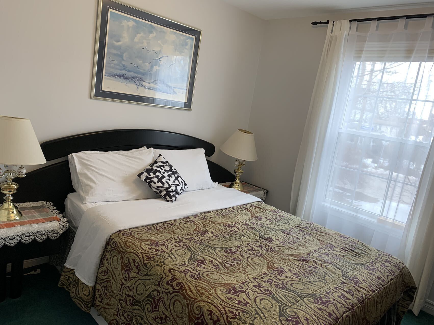 A neatly made bed with a patterned bedspread, two bedside tables with lamps, a framed artwork above the bed, and a window with sheer curtains.