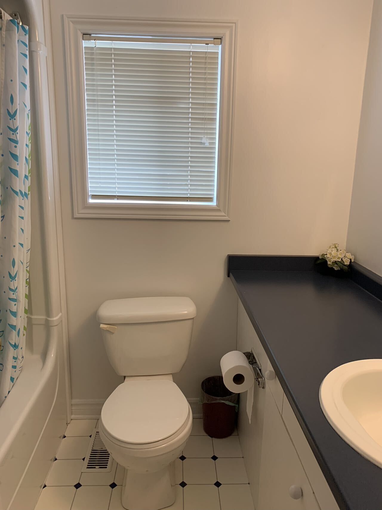 A bathroom with a toilet, a window with blinds, a shower curtain, a sink, and a trash can.